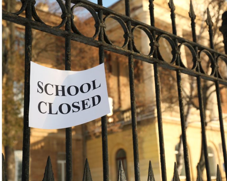 A piece of paper with the phrase "SCHOOL CLOSED" attached to a metal fence with a large building behind it.