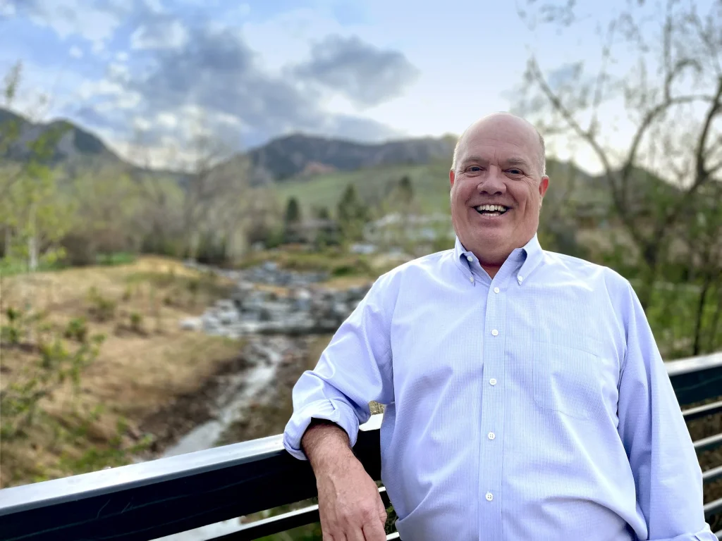 A photo of a man in a light blue button-down shirt leaning against the railing of a bridge. In the background there are mountains, trees, and a small creek.