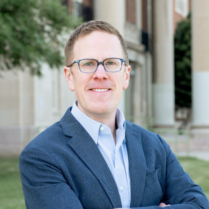 A headshot of a man with short dirty blond hair, blue glasses, and a dark blue suit jacket.